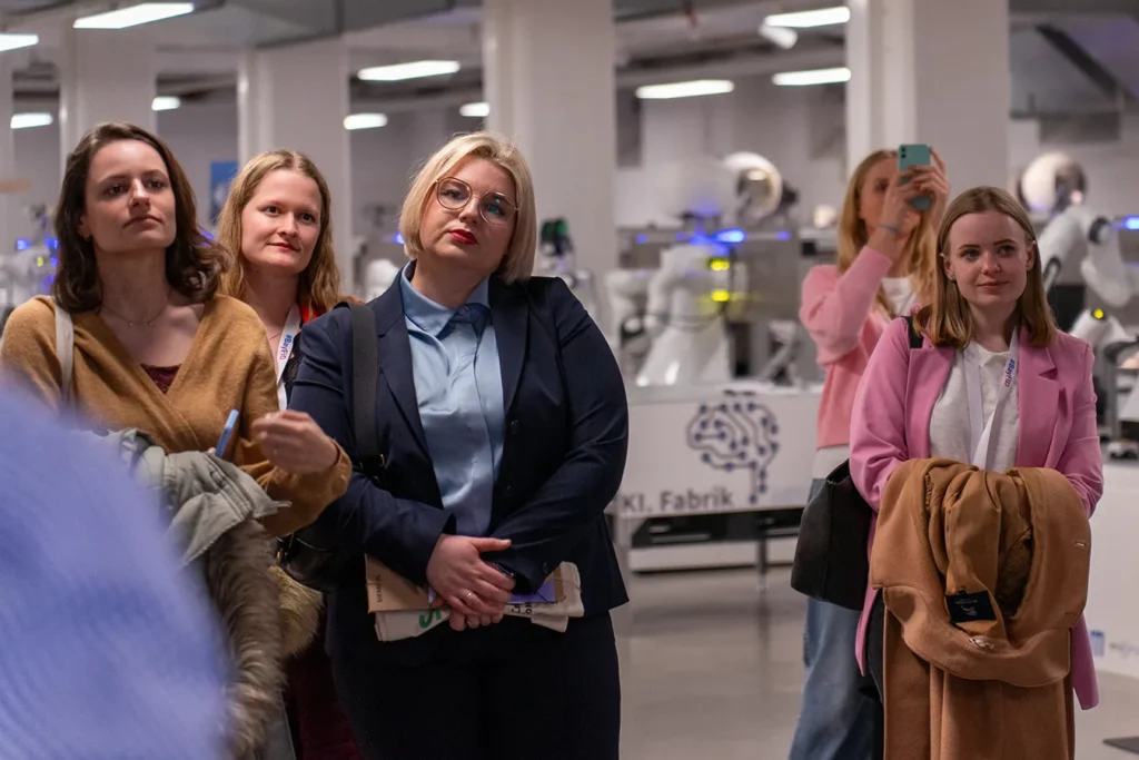 5 Frauen stehen in der KI-Fabrik und blicken interessiert, im Hintergrund sind Roboterarme zu sehen